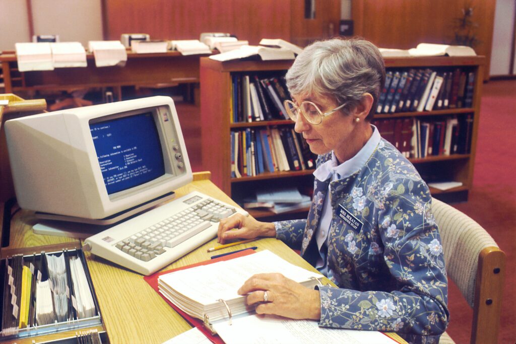 Older woman at a desk with folder, pens and IBM computer.